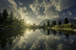 Riflesso perfetto di sole starburst e pini su un lago alpino cristallino delle Dolomiti all'alba, composizione di Carlo Pagliuca