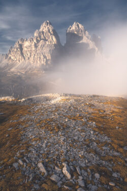 Dettaglio del terreno detritico in primo piano con le Tre Cime di Lavaredo che emergono dalla nebbia, scatto tecnico di paesaggio