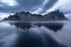 Le iconiche cime Vestrahorn riflesse sulla spiaggia nera bagnata sotto un cielo scuro e drammatico, fotografia di paesaggio islandese