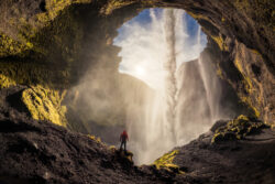 Escursionista in giacca rossa osserva la cascata Kvernufoss dall'interno della grotta in Islanda, fotografia di paesaggio di Carlo Pagliuca